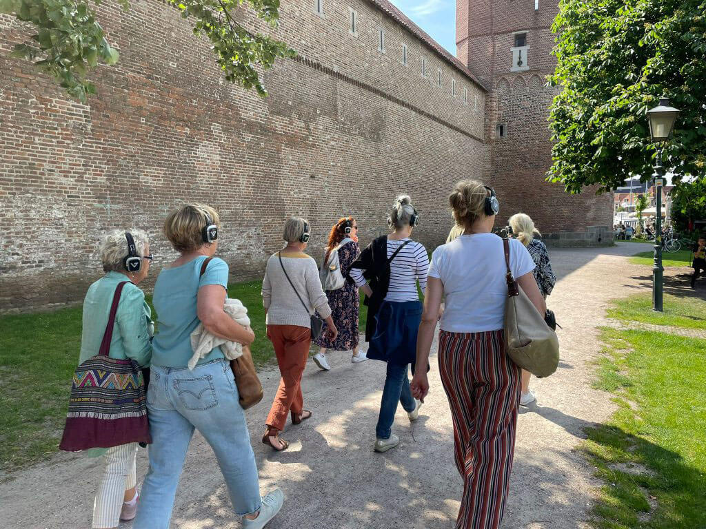 Groep wandelende mensen, van achter gefotografeerd, luistert een audioverhaal via een koptelefoon.